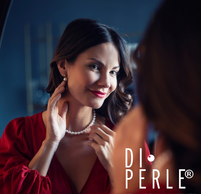 Eine Frau in einem roten Kleid und einer Perlenkette schaut in einen Spiegel und berührt ihre Kette. Das Bild enthält ein Wasserzeichen mit der Aufschrift 'DI PERLE'.