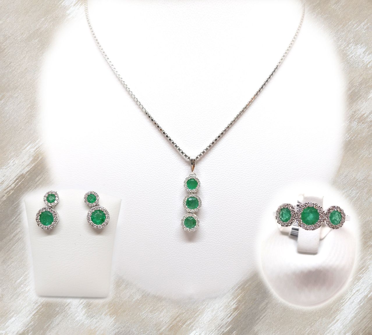 Eine Silberkette mit Anhänger und drei Smaragden, begleitet von passenden Ohrringen und einem Ring, auf weißem Hintergrund präsentiert.