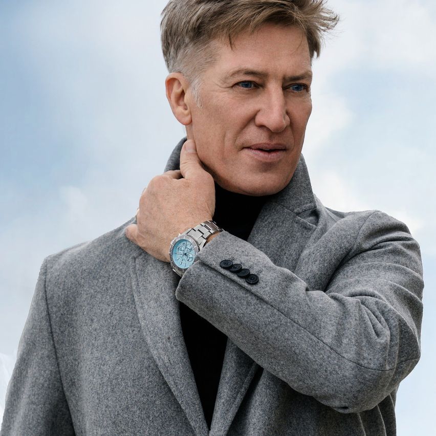 Ein Mann in einem grauen Mantel und schwarzem Rollkragenpullover berührt seinen Hals mit der linken Hand und trägt eine blaue Uhr am linken Handgelenk.