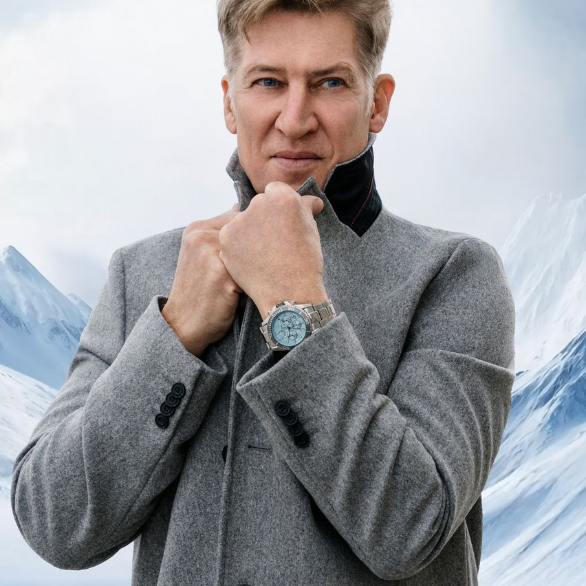 Ein Mann in einem grauen Mantel justiert seinen Kragen und trägt eine silberne Uhr am linken Handgelenk. Er steht vor einer verschneiten Berglandschaft.