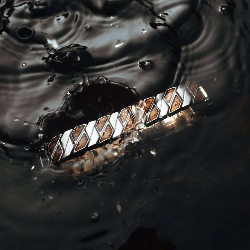 Ein metallisches Armband mit abwechselnden silbernen und braunen Mustern ist in dunklem Wasser mit Wellen untergetaucht.