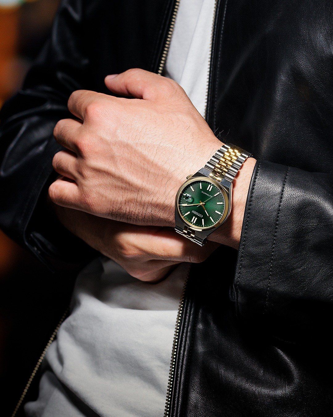 Ein Mann in einer schwarzen Lederjacke und einem weißen T-Shirt hält eine Uhr mit grünem Zifferblatt.