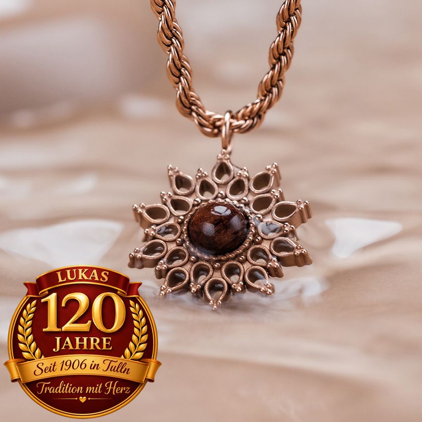 Eine kupferne Halskette mit einem braunen Steinanhänger in Form einer Blume liegt auf einem beigefarbenen Tuch. Der Anhänger ist an einer Kupferkette befestigt. Das Bild ist mit Lukas 120 Jahre und einem Lorbeerkranz darunter gekennzeichnet.