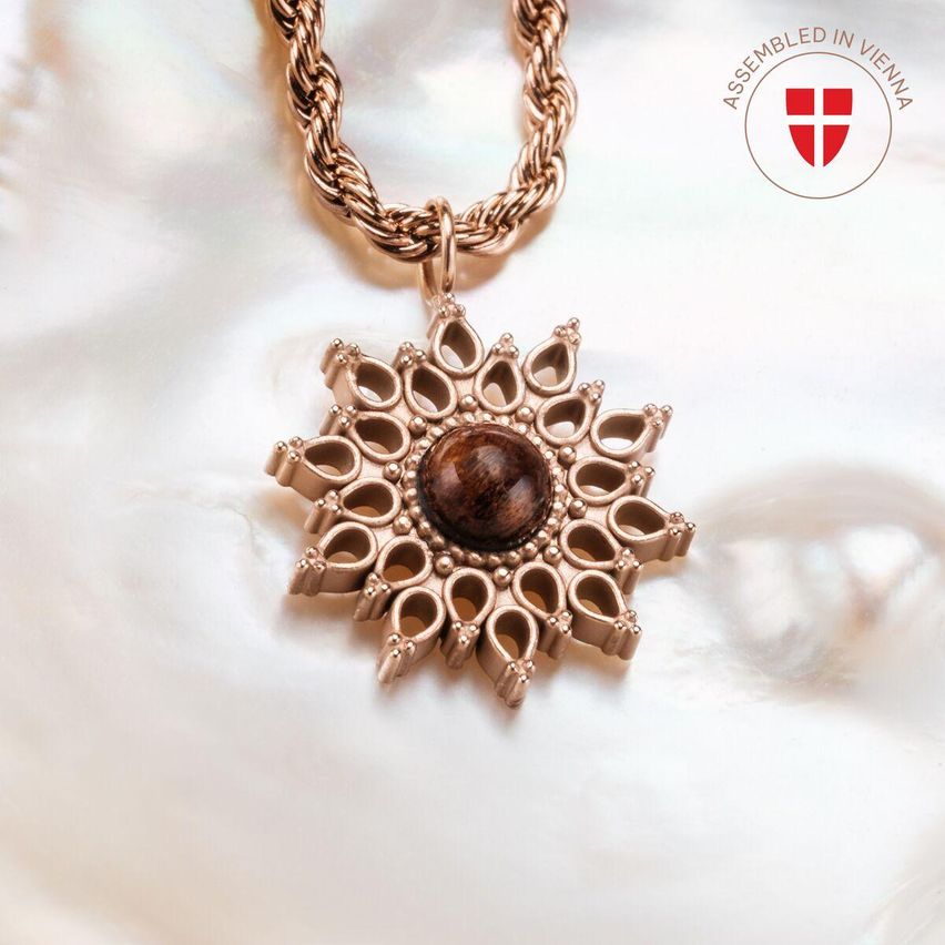 Eine kupferne Halskette mit einem blumenförmigen Anhänger, der einen braunen Stein enthält, liegt auf einer weißen Oberfläche. Sie wird in Wien zusammengesetzt.