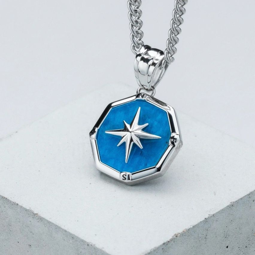 Eine Silberkette mit einem blauen sechseckigen Anhänger, der einen weißen Stern und die Buchstaben SI zeigt. Der Anhänger ist auf einer weißen Oberfläche ausgestellt.