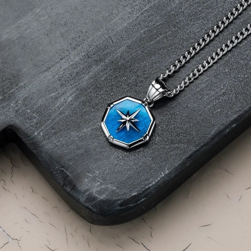 Eine Silberkette mit einem sechseckigen Anhänger, mit blauem Zentrum und einem weißen Stern, auf einer grauen Oberfläche liegend.