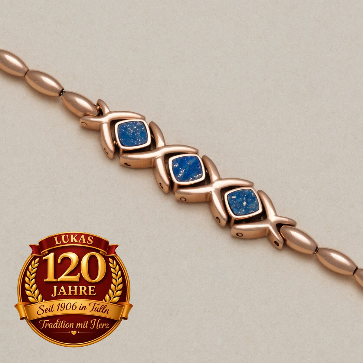 Ein Armband mit Roségold-Design und blauen Edelsteinen liegt auf einem beigefarbenen Hintergrund. Unten ist ein Logo für Lukas zu sehen, das 120 Jahre feiert.