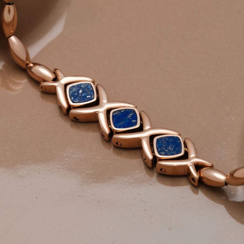 Ein Armband aus Roségold mit blauen Steinen liegt auf einer braunen Oberfläche. Es hat vier blaue Steine und ein einzigartiges Design.