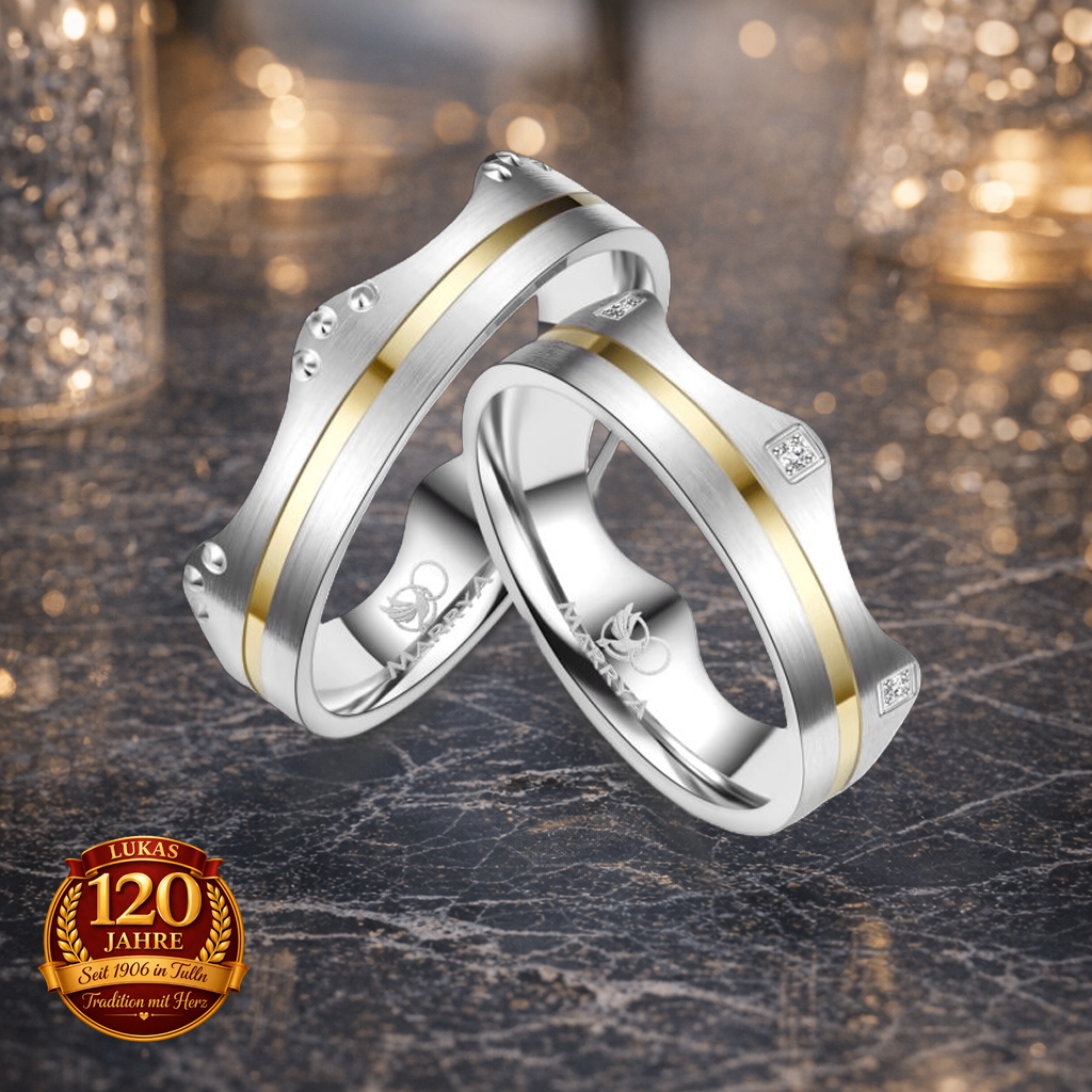 Zwei Silberringe mit Goldakzenten und Diamanten ruhen auf einer Marmoroberfläche und tragen ein Logo und 'Lukas 120 Jahre' Branding.