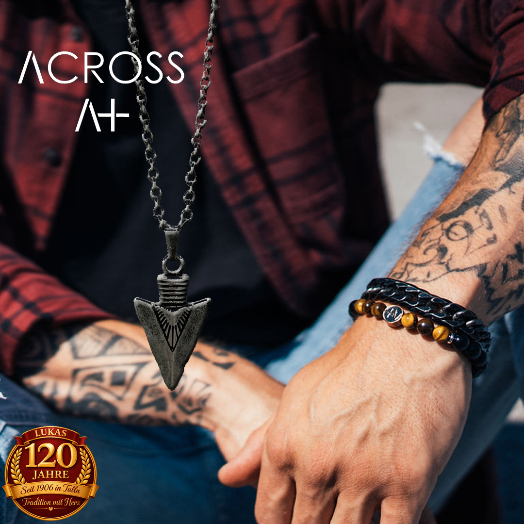Ein Mann mit einem roten Hemd und Jeans, mit einer silbernen Pfeilspitzen-Halskette, sitzt mit einer Tätowierung am Arm. Ein schwarzes Armband mit braunen und gelben Perlen ist am anderen Arm.
