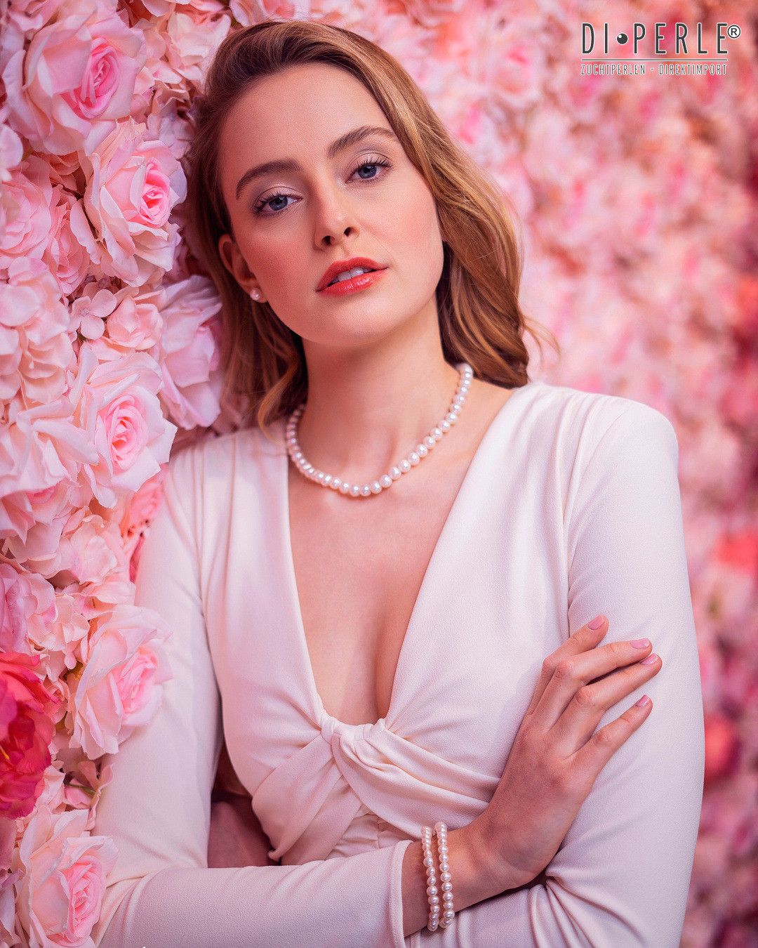 Eine Frau mit blondem Haar, die ein weißes Oberteil und eine Perlenkette trägt, posiert vor einem Hintergrund aus rosa Rosen.