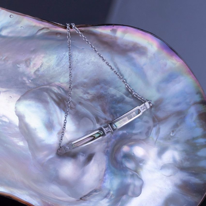 Eine Silberkette mit grünen und weißen Steinen ruht auf einer Muschel mit regenbogenartigem Farbton. Die Kette hat ein stabförmiges Design mit Diamanten.