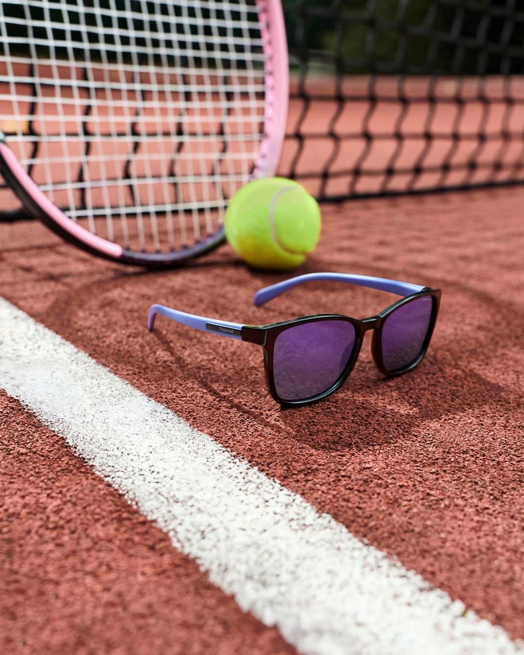 Ein Paar Sonnenbrille mit lila Gläsern liegt auf einem Tennisplatz neben einem Tennisball und einem Tennisschläger.