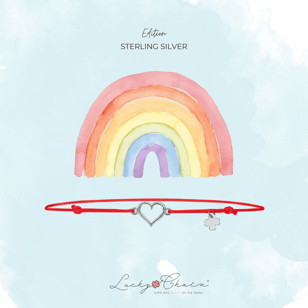Eine farbenfrohe Regenbogenillustration auf blauem Hintergrund mit einem roten Armband, das ein Herz-Anhänger und ein kleines Kreuz-Anhänger hat. Der Text 'Edition STERLING SILVER' ist über dem Regenbogen angezeigt.