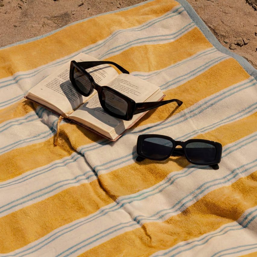 Ein Paar schwarze Sonnenbrille liegt auf einem offenen Buch, das auf einem gelb-blau gestreiften Handtuch am Strand liegt.