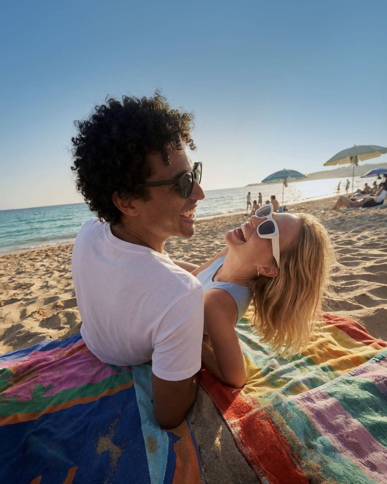 Ein Mann und eine Frau sitzen am Strand, lächeln und schauen in die Kamera. Sie tragen Sonnenbrillen und sitzen auf einer bunten Decke.