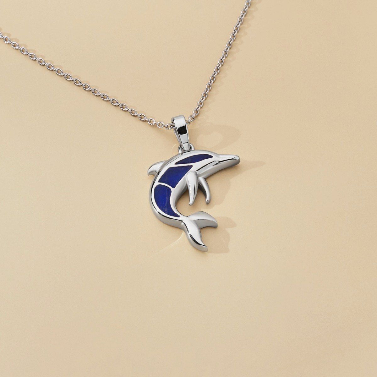 Ein silberner Delfin-Anhänger mit blauer Emaille auf einer silbernen Kette, auf einem beigefarbenen Hintergrund präsentiert.
