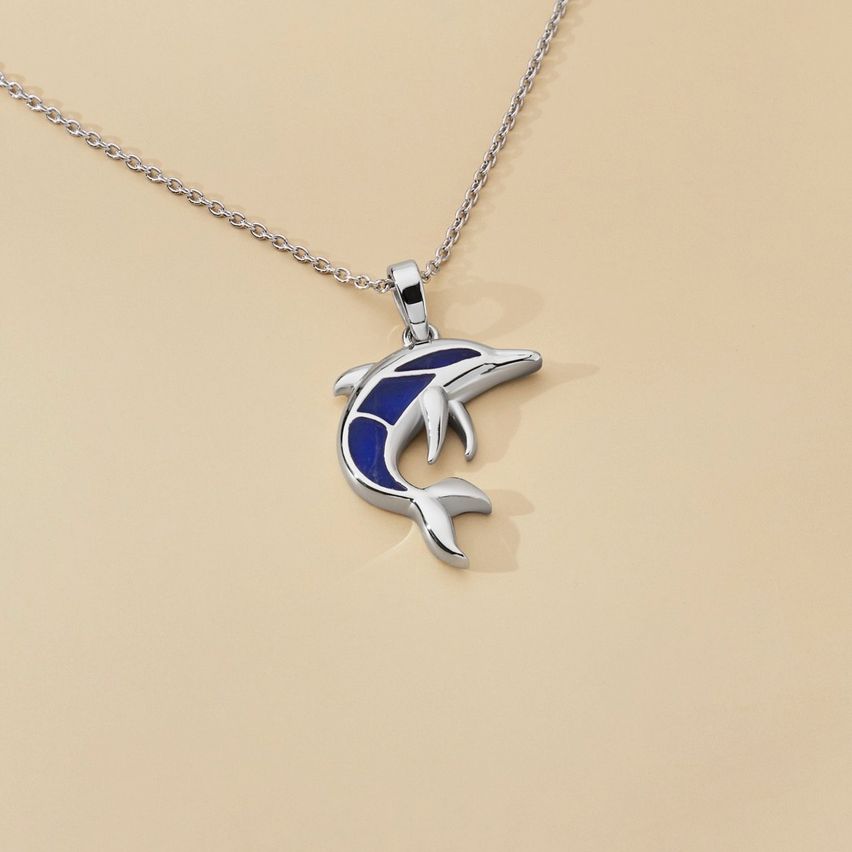 Ein silberner Delfin-Anhänger mit blauer Emaille auf einer silbernen Kette, auf einem beigefarbenen Hintergrund präsentiert.