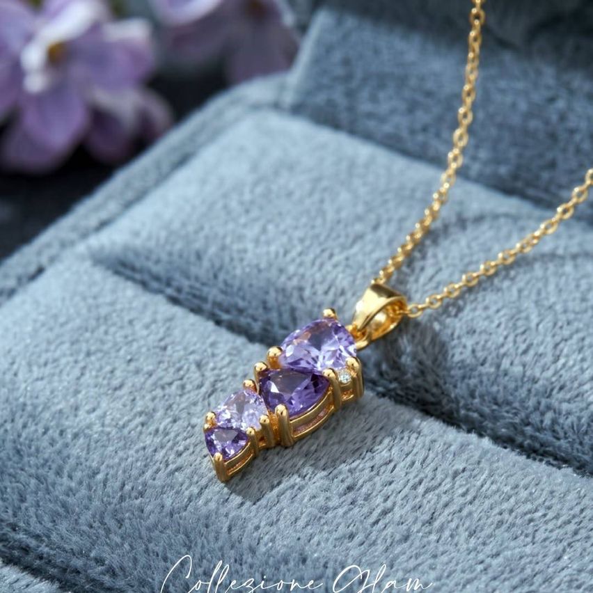 Eine goldene Halskette mit einem Anhänger, der drei lila Edelsteine aufweist, auf einem blauen Samttuch.