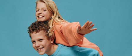 Zwei Kinder, ein Junge und ein Mädchen, stehen vor einem blauen Hintergrund. Das Mädchen sitzt auf den Schultern des Jungen, beide lächeln.