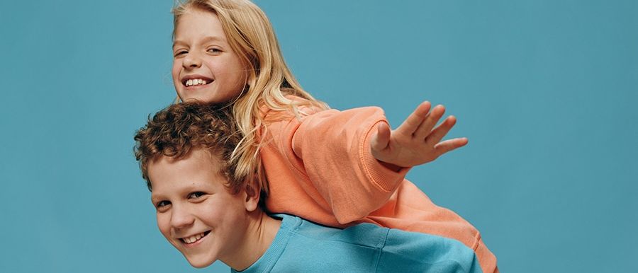 Zwei Kinder, ein Junge und ein Mädchen, stehen vor einem blauen Hintergrund. Das Mädchen sitzt auf den Schultern des Jungen, beide lächeln.