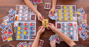 Kinder spielen ein Kartenspiel auf einem Holztisch, umgeben von verschiedenen Karten mit verschiedenen Ländern und einer goldenen Trophäenkarte in der Mitte.