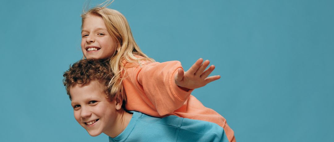 Ein junger Junge mit lockigen Haaren trägt ein lachende Mädchen auf seinen Schultern. Das Mädchen trägt ein orangefarbenes Sweatshirt, und der Junge trägt ein blaues Sweatshirt. Sie lächeln beide und scheinen für ein Foto zu posieren.