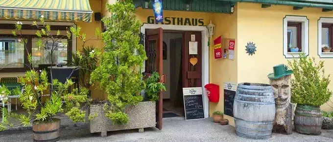Der Eingang zu einem gelben Gebäude mit einem grünen Vordach und einem Schild, das Gasthaus liest. Draußen stehen Topfpflanzen und Fässer.