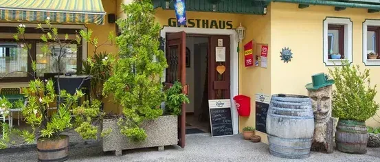 The entrance to a yellow building with a green awning and a sign reading Gasthaus. There are potted plants and barrels outside.