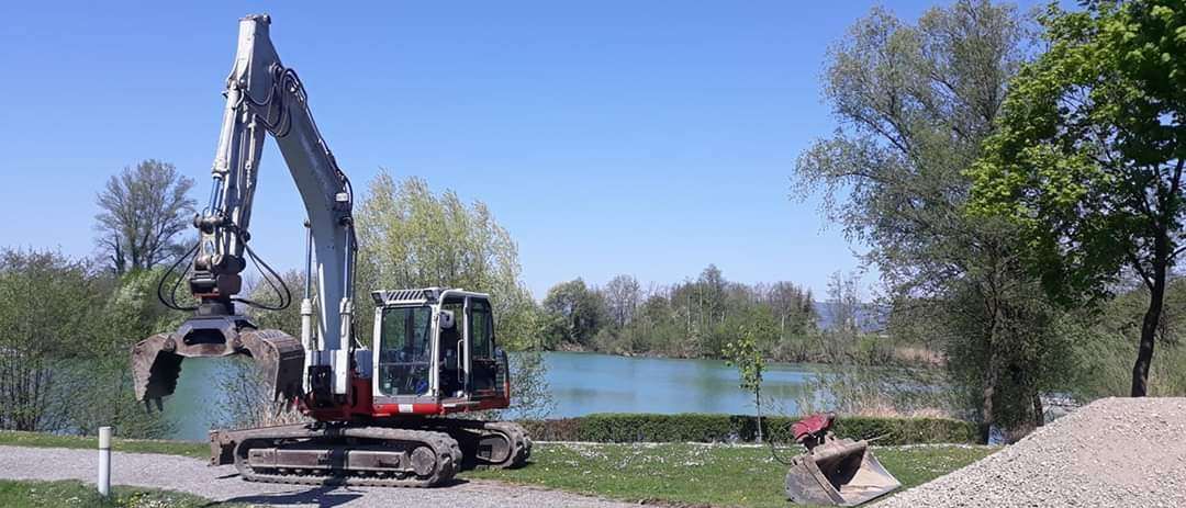 Ein Bagger steht auf einem Kiesweg in der Nähe eines Sees mit Bäumen und Büschen. Der See ist ruhig und spiegelt den klaren Himmel wider.