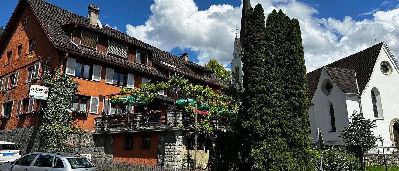 Ein silbernes Auto fährt an einem Gebäude mit Balkon und grünen Schirmen vorbei, in der Nähe einer Kirche und eines großen grünen Baumes.