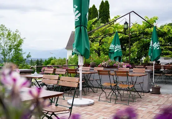 Ein Outdoor-Restaurant mit grünen Sonnenschirmen, Holztischen und Stühlen, umgeben von Bäumen und Blumen, mit Blick auf eine entfernte Bergkette.