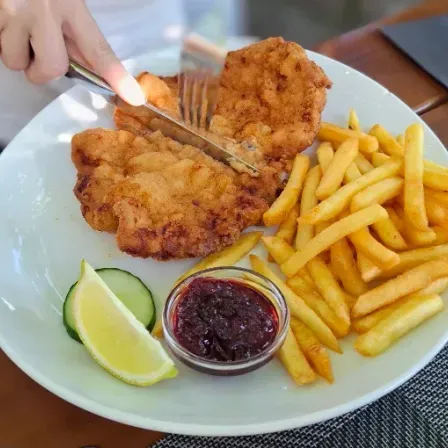 Ein weißer Teller mit gebratenem Fisch, Pommes Frites, einer Zitronenscheibe und einer kleinen Schüssel mit Marmelade. Eine Person schneidet den Fisch mit Messer und Gabel.