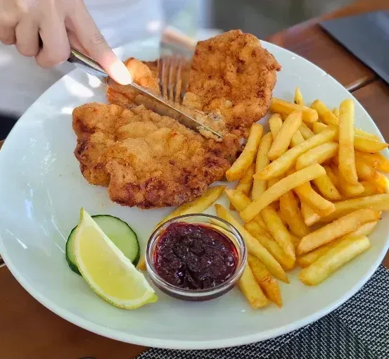 Ein weißer Teller mit gebratenem Fisch, Pommes Frites, einer Zitronenscheibe und einer kleinen Schüssel mit Marmelade. Eine Person schneidet den Fisch mit Messer und Gabel.