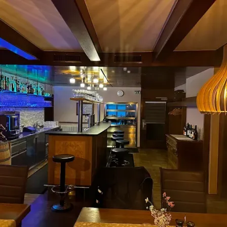 Das Innere einer Bar mit einer hölzernen Bartheke, Stühlen und einem Fass mit Weinflaschen. Eine große Lampe hängt von der Decke.