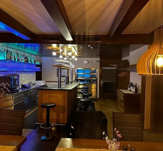 Das Innere einer Bar mit einer hölzernen Bartheke, Stühlen und einem Fass mit Weinflaschen. Eine große Lampe hängt von der Decke.