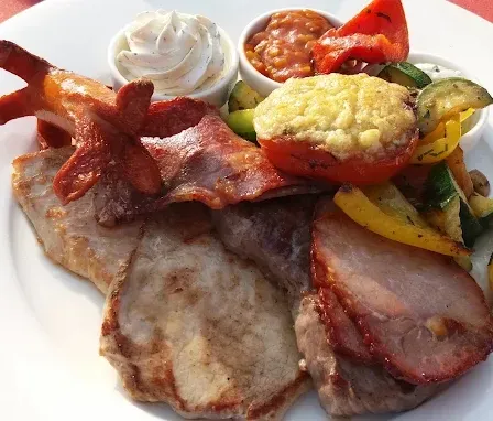 Ein weißer Teller mit verschiedenen Fleischsorten, Speck und Gemüse wie Tomaten, Paprika und Zucchini, begleitet von zwei kleinen Schüsseln mit Beilagen.