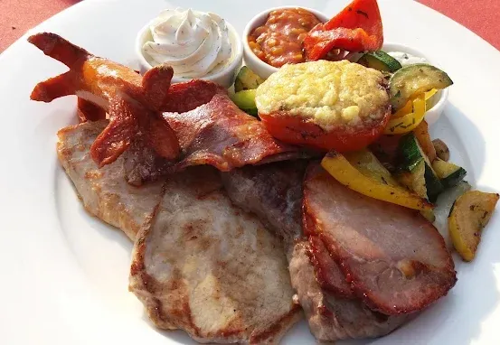 Ein weißer Teller mit verschiedenen Fleischsorten, Speck und Gemüse wie Tomaten, Paprika und Zucchini, begleitet von zwei kleinen Schüsseln mit Beilagen.