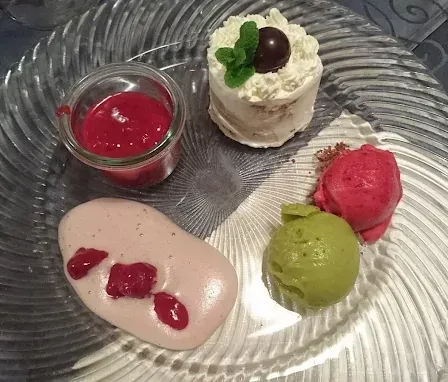 Ein Glasteller mit verschiedenen Desserts: ein kleiner Kuchen mit weißer Glasur und einem grünen Minzblatt, eine Kugel rosa Sorbet, eine Kugel grünes Sorbet und ein rosa Dessert mit roten Beeren.