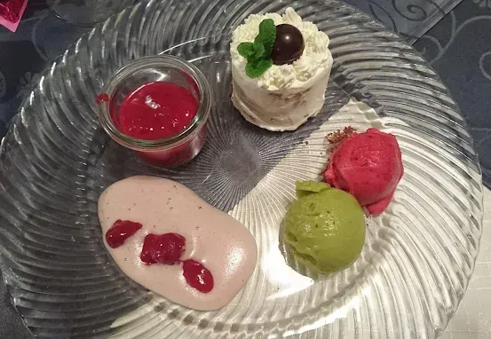 Ein Glasteller mit verschiedenen Desserts: ein kleiner Kuchen mit weißer Glasur und einem grünen Minzblatt, eine Kugel rosa Sorbet, eine Kugel grünes Sorbet und ein rosa Dessert mit roten Beeren.