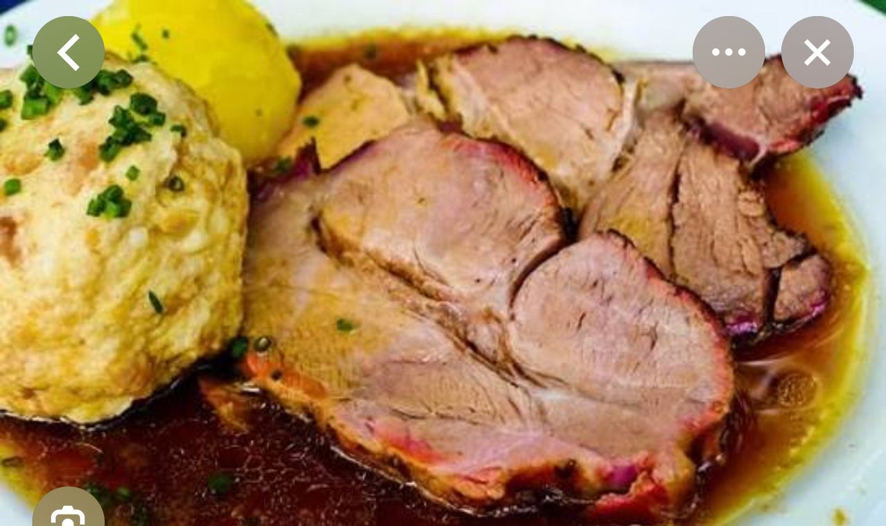 Ein Nahaufnahme eines Gerichts mit reichhaltiger Sauce, bestehend aus Schweinebratenscheiben, einer goldenen Kartoffel und einer Spur Kräutergarnitur.