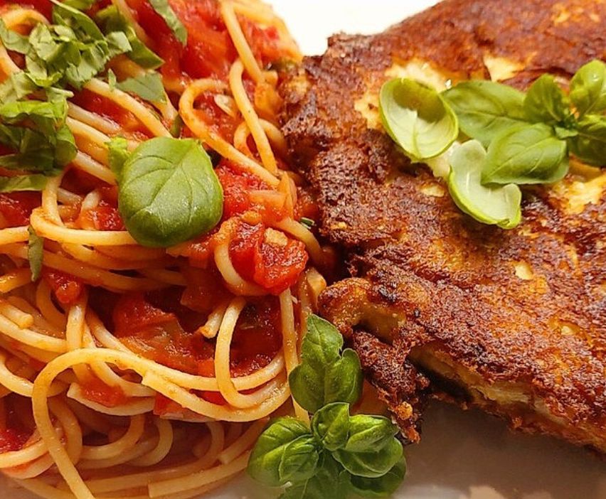 Ein Teller mit Spaghetti in Tomatensoße und Basilikumblättern, neben einem knusprigen panierten Hähnchenschnitzel garniert mit Basilikum.
