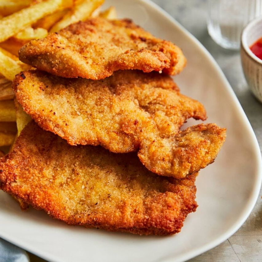 Ein Teller mit drei Stücken panierter Hähnchenbrust, goldbraun und knusprig, begleitet von einer Beilage goldener Pommes Frites, serviert mit einem kleinen Schälchen Ketchup auf der Seite.