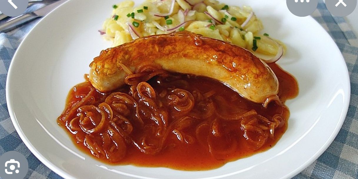 Ein Teller mit einer gekochten Wurst, überzogen mit karamellisierten Zwiebeln und brauner Sauce, begleitet von Kartoffelpüree und geschnittenen Zwiebeln.