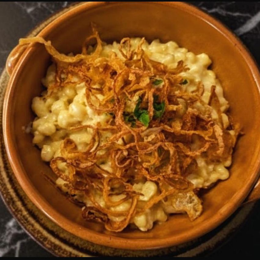 Eine Schüssel mit Makkaroni und Käse, garniert mit knusprigen gebratenen Zwiebeln und grünen Kräutern auf einer Marmoroberfläche.