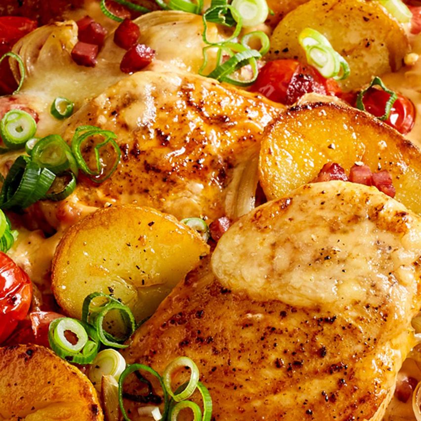 Eine Nahaufnahme eines Gerichts mit gebratenem Hähnchen, goldenen Kartoffeln und einer Mischung aus grünen und roten Gemüsen, überzogen mit einer cremigen Sauce.