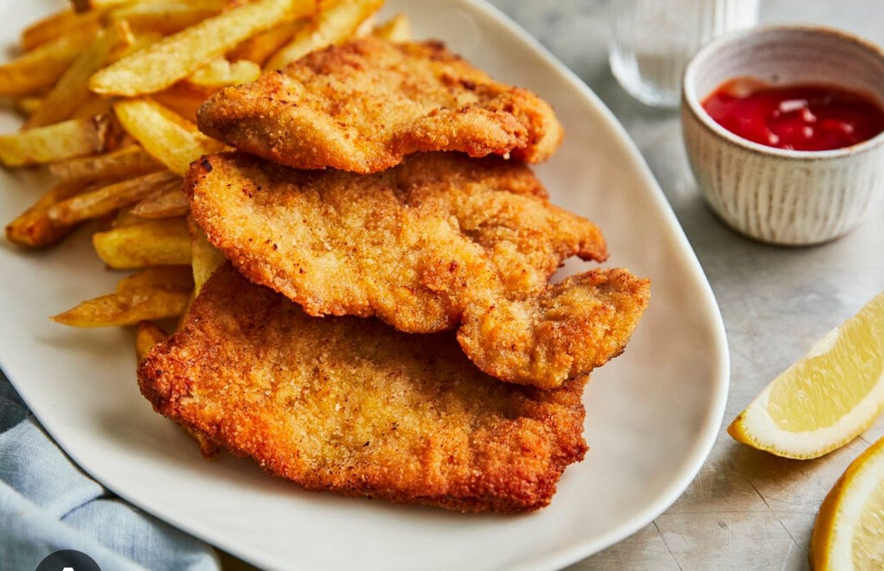 A white plate displays a stack of golden-brown breaded chicken cutlets, with French fries on the side. A bowl of ketchup is nearby.