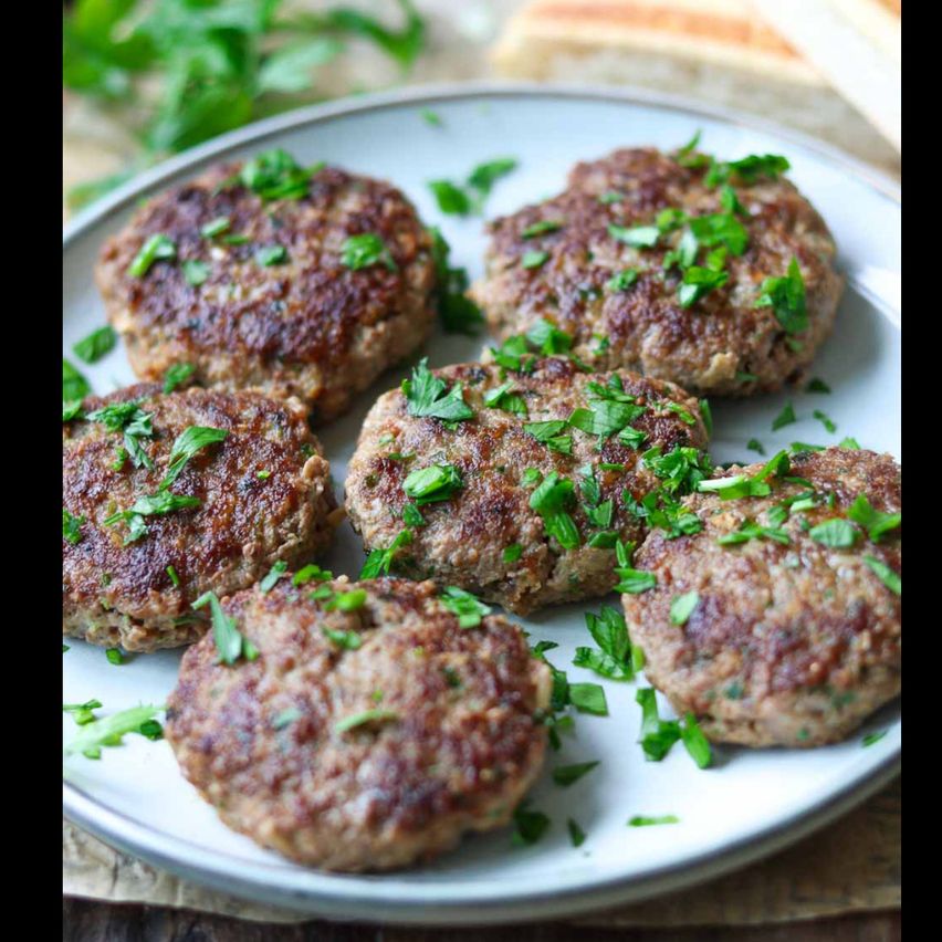 A white plate holds six cooked meat patties garnished with chopped parsley, placed on a wooden surface.