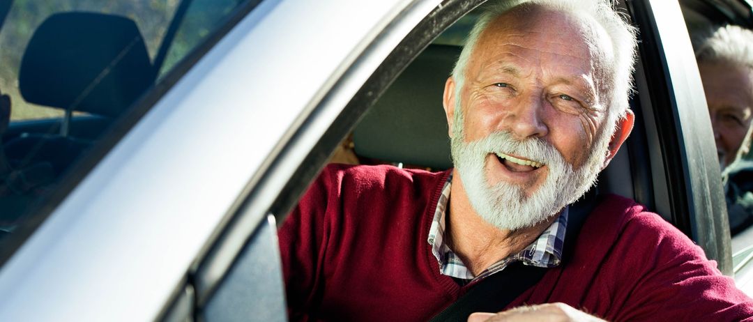 Ein älterer Mann lächelt, während er in einem Auto mit einem weißen Logo namens 'emilf' sitzt. Er trägt einen roten Pullover und einen weißen Bart.