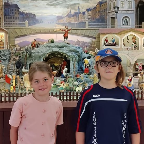 Zwei junge Mädchen stehen vor einer detaillierten Krippenszene mit Figuren und einem Hintergrund einer Stadt.